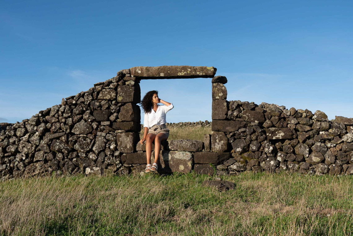 Woman sitting on a traditional basalt stone wall with a rectangular opening, surrounded by grasslands and clear skies in rural Terceira Island, Azores.