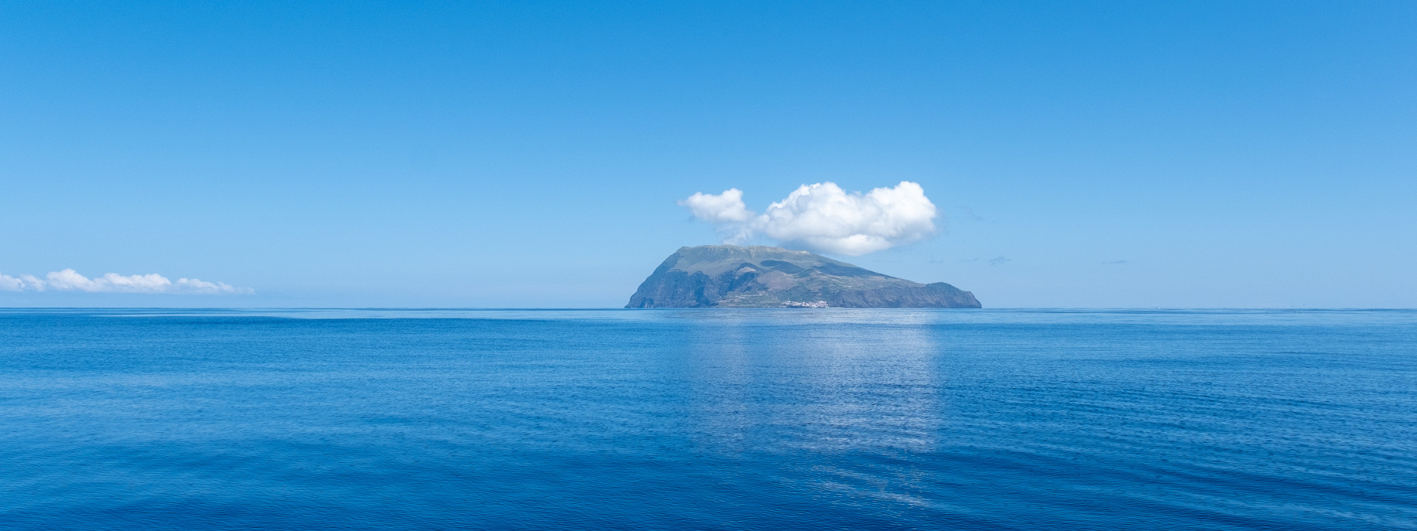 Corvo Island rises from the calm Atlantic Ocean in the Azores, Portugal. Its volcanic slopes and coastal settlement are visible beneath a small bank of white clouds.