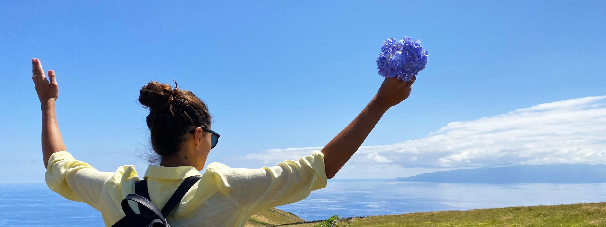 A hiker holding blue hydrangeas looks across the Atlantic from the highlands of Corvo Island in the Azores, Portugal. Open grassland and distant ocean views define the island’s elevated volcanic landscape.