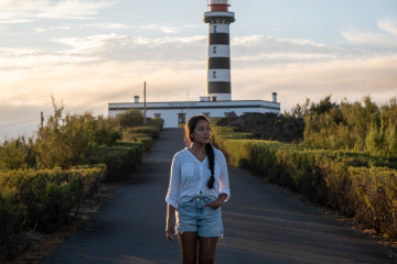 Traveler walking toward Ponta da Barca Lighthouse on Graciosa Island, with its black-and-white striped tower and red lantern overlooking the Atlantic coast. The 23-meter tower is the tallest lighthouse tower in the Azores.