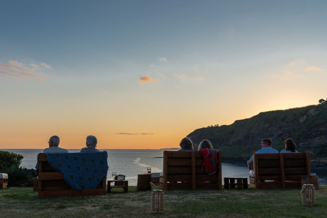Guests enjoying a peaceful sunset overlooking the ocean from wooden lounge chairs at Caloura Hotel in São Miguel Island, Azores.
