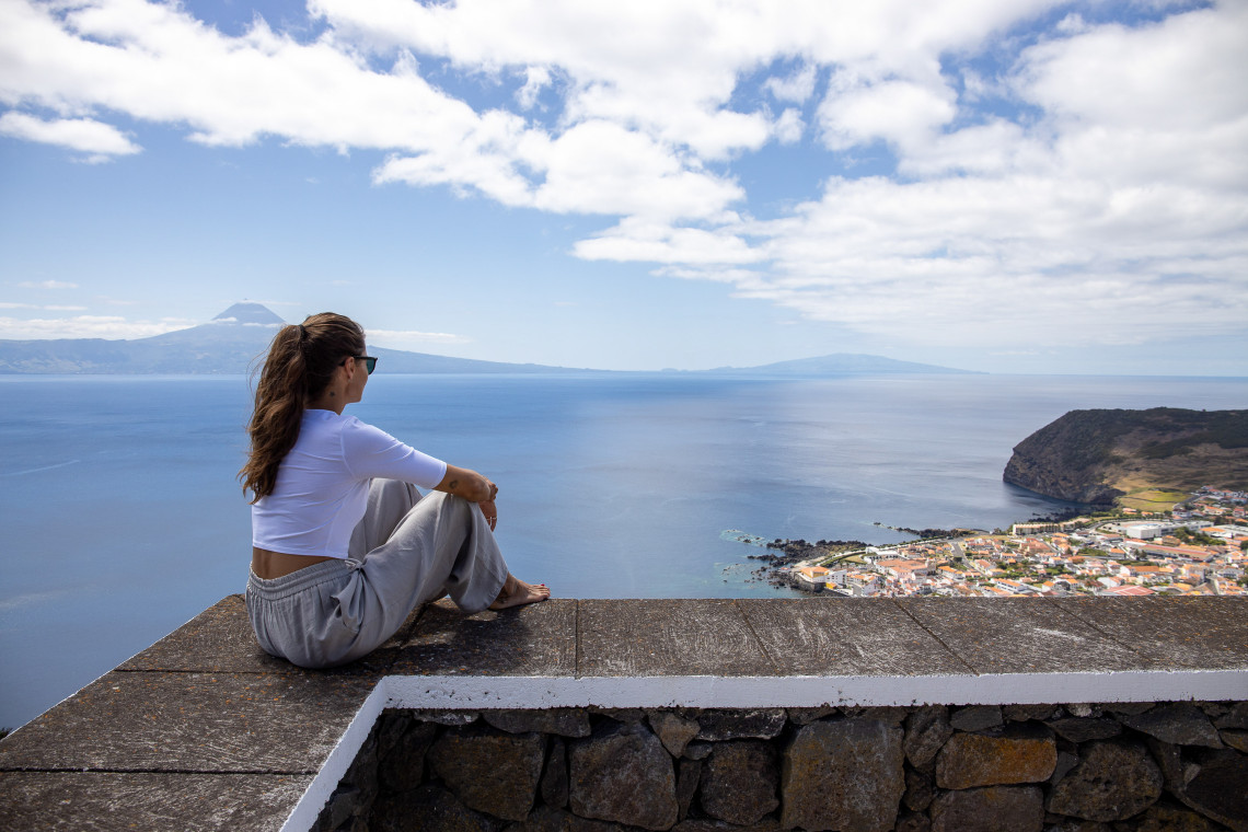 Woman admiring Pico Mountain from Faial Island, Azores Woman sitting on a viewpoint in Faial Island, Azores, looking at Pico Mountain across the sea under a blue sky with scattered clouds.