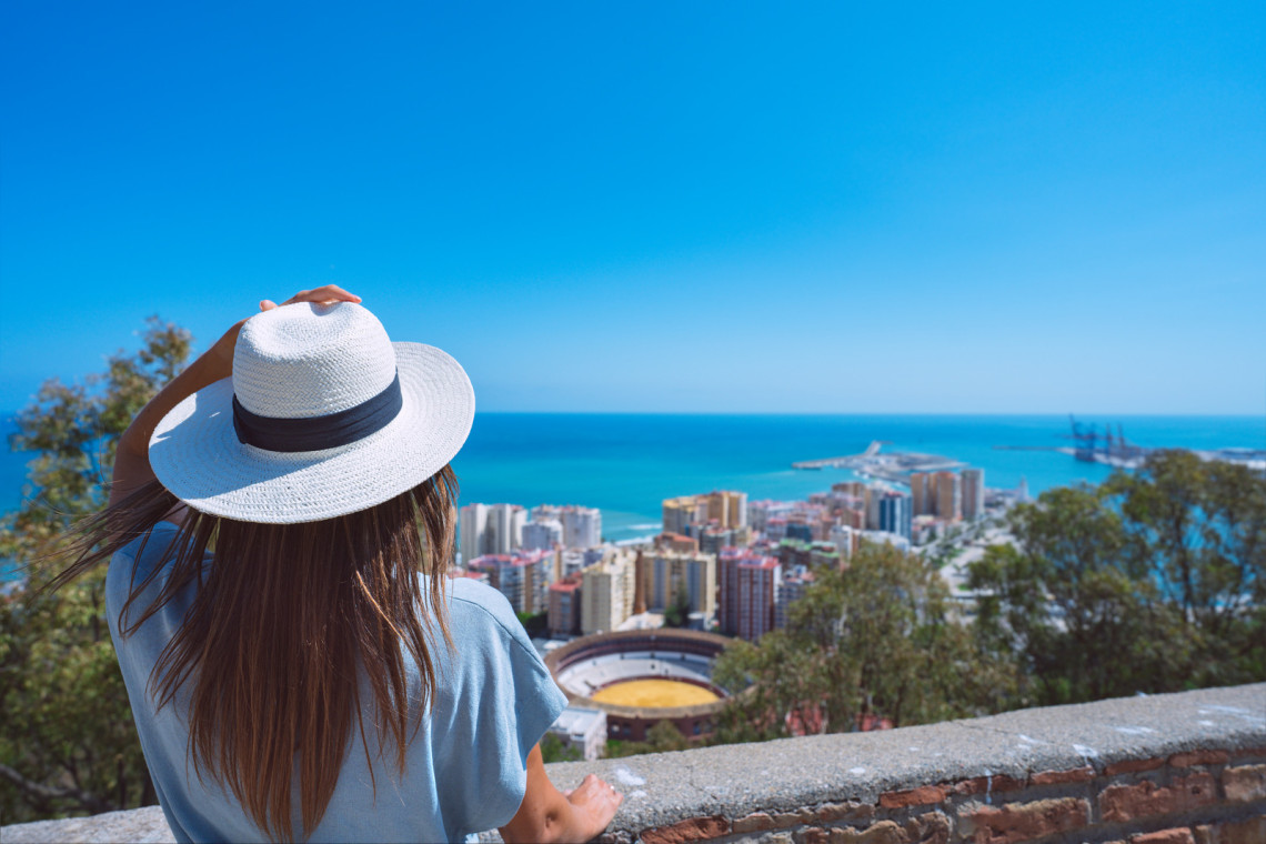 Rear view of young woman traveller with white hat standing in Gibralfaro Castle viewpoint and looking at Malaga cityscape. Summer holiday vacation in Spain rear-view-of-young-woman-traveller-with-white-hat-standing-in-gibralfaro-castle-viewpoint-and-looking-at-malaga-cityscape-summer-holiday-vacation-in-spain