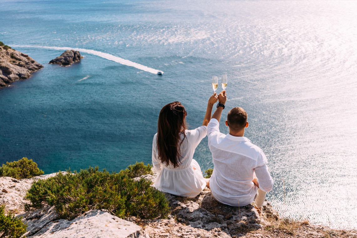 Couple sitting on a cliff in the Algarve, Portugal, raising champagne glasses while overlooking the Atlantic Ocean.