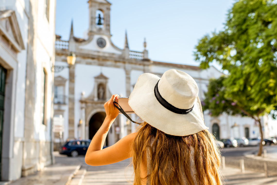 Woman wearing a white hat taking a photo of Arco da Vila in Faro’s Old Town, Algarve, Portugal.