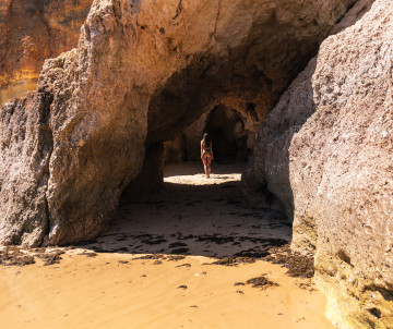 A person walking through a natural rock cave opening on Praia do Vau beach in Algarve, Portugal, with golden sand and rugged cliffs.