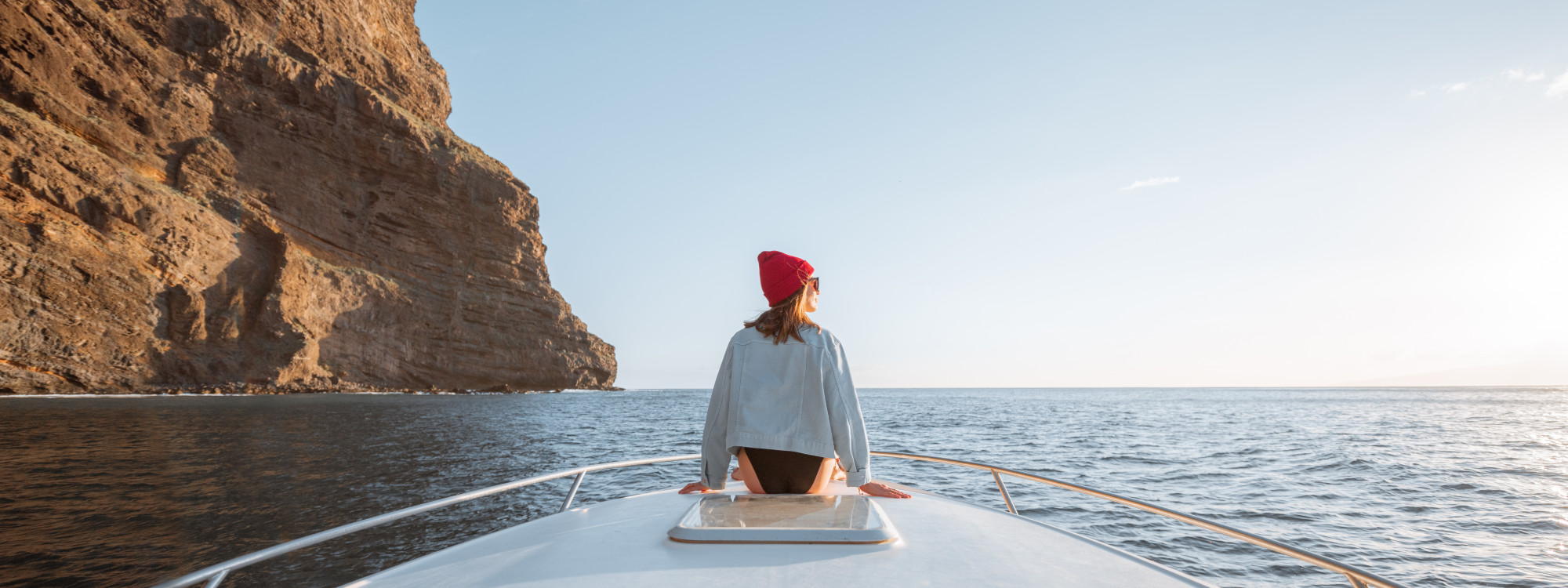 Coastal Boat Tour Along the Algarve Cliffs Person sitting at the front of a boat, wearing a red beanie, looking toward the sea and towering cliffs during a boat tour along the Algarve coast in Portugal