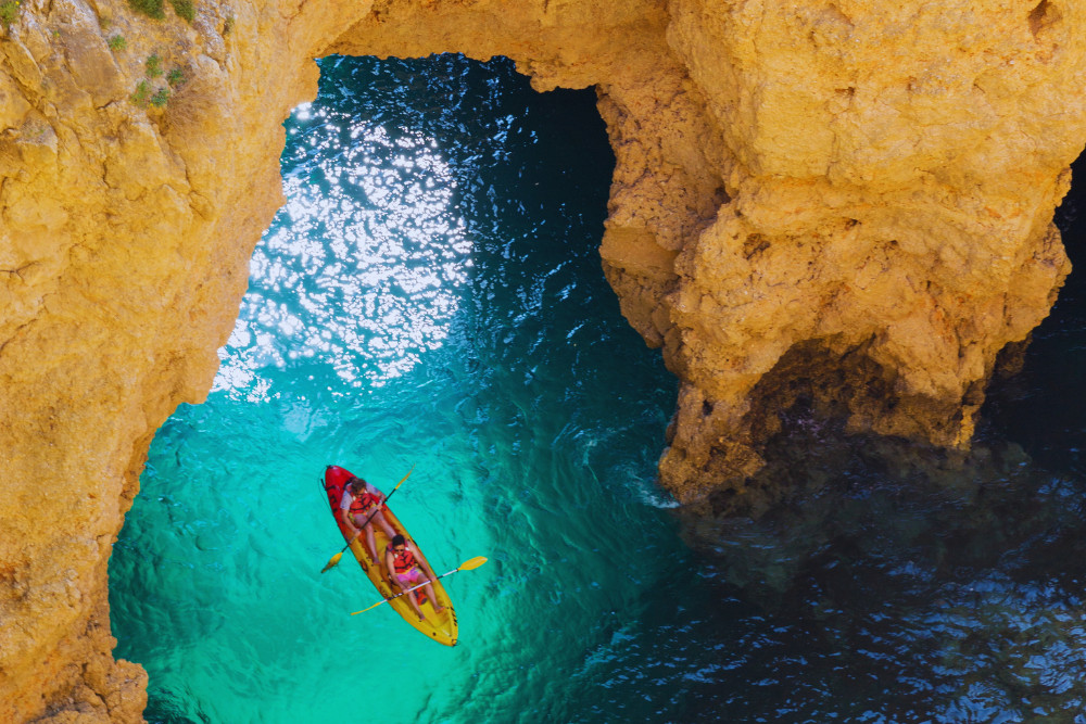 Aerial view of two people kayaking through turquoise waters between dramatic limestone cliffs in the Algarve, Portugal, on a coastal cave exploration adventure