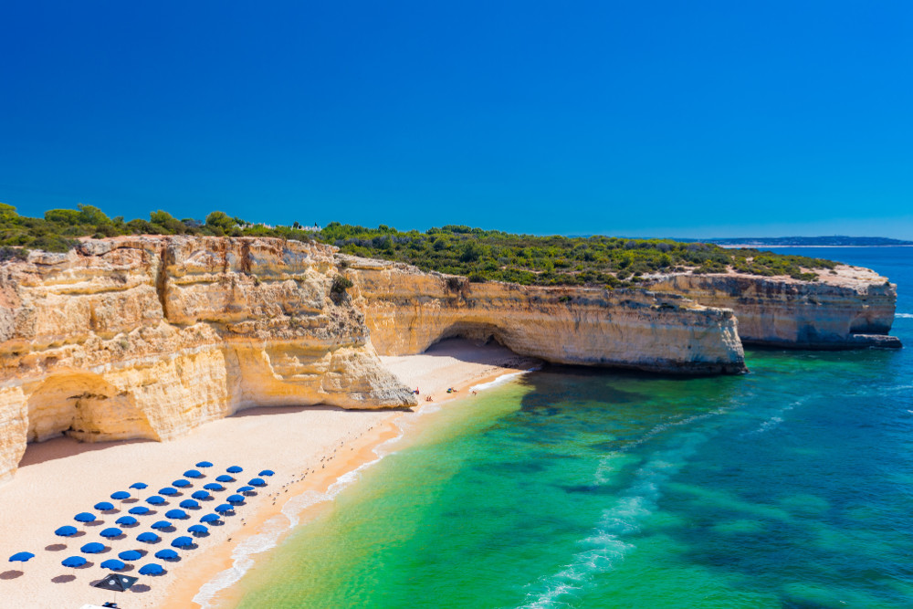 Aerial view of Praia da Marinha in Algarve, Portugal, with golden limestone cliffs, turquoise sea, and rows of blue umbrellas on the sandy beach