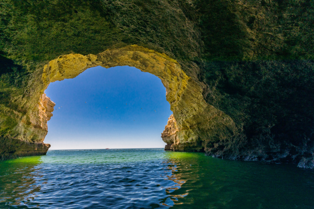 View from inside a sea cave along the Algarve coastline in Portugal, showing clear greenish-blue water and sunlight framing the cave entrance toward the Atlantic Ocean