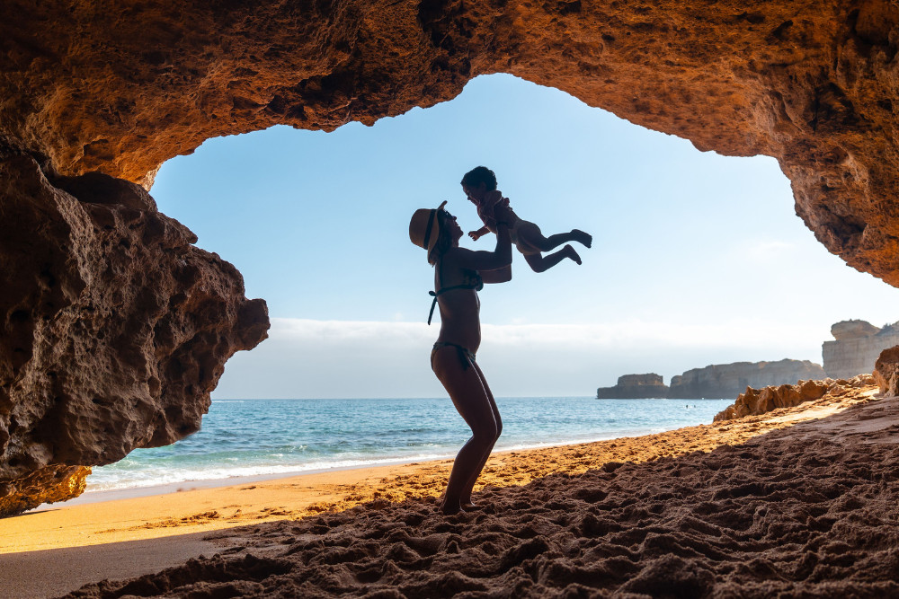 Mother lifting her child beneath a natural rock formation on Coelha Beach in the Algarve, Portugal, with golden sand, sea cliffs, and the Atlantic Ocean in the background