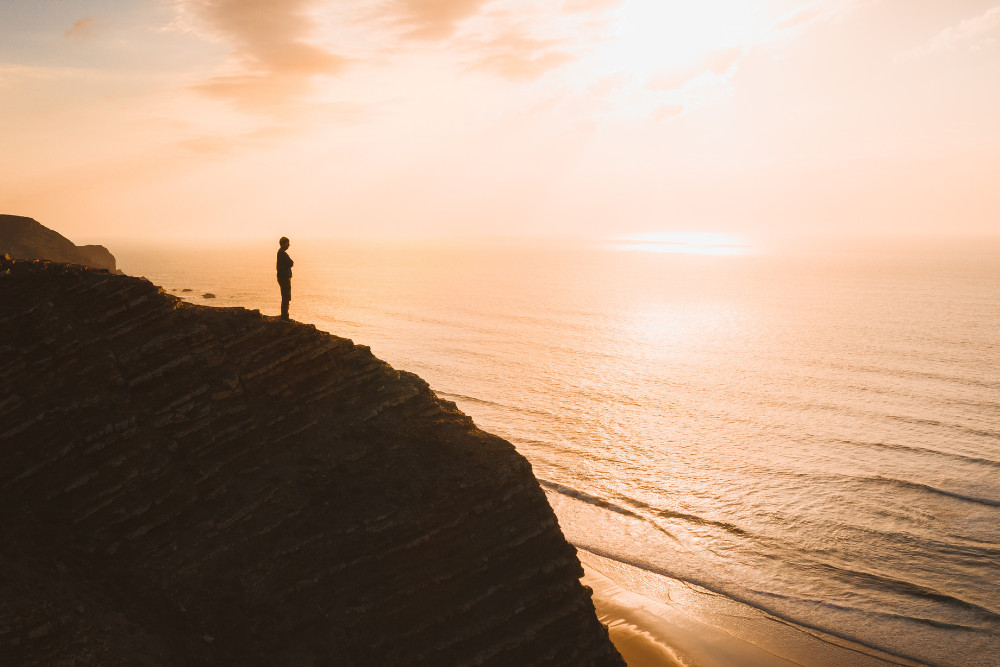 Silhouette of a person standing on a high cliff at sunset, overlooking the Atlantic Ocean and a quiet beach along the Algarve coastline in Portugal
