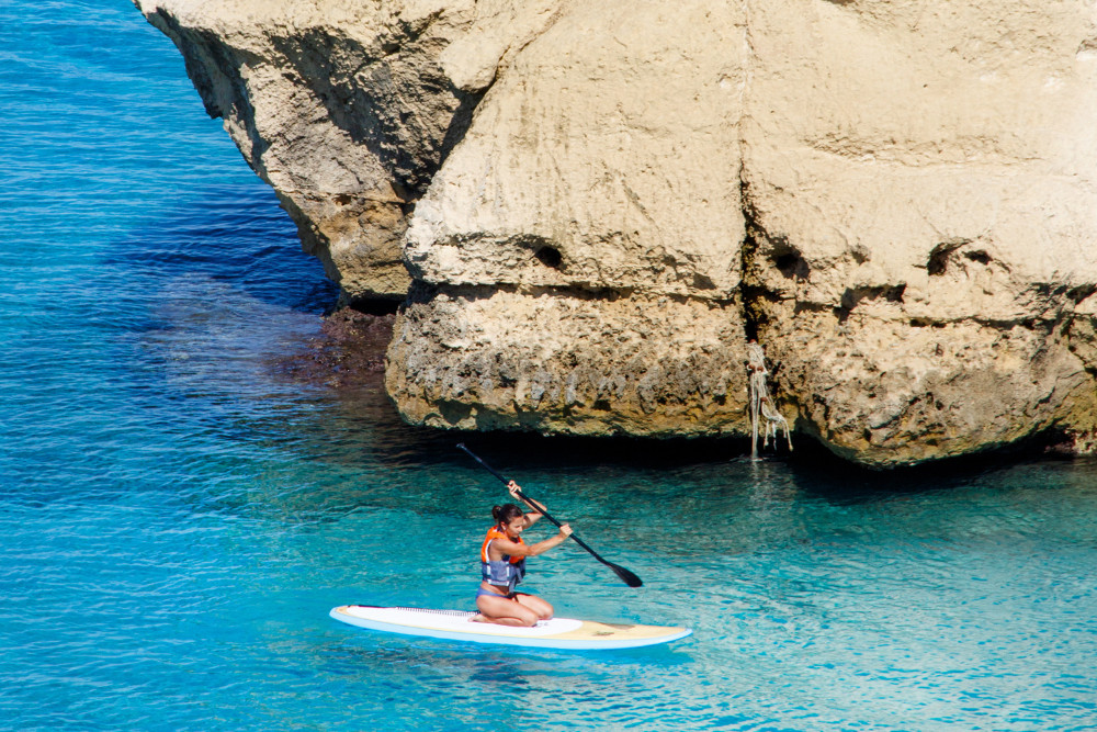 Woman paddleboarding in clear turquoise waters along the rocky coastline of the Algarve, Portugal, near dramatic limestone cliffs