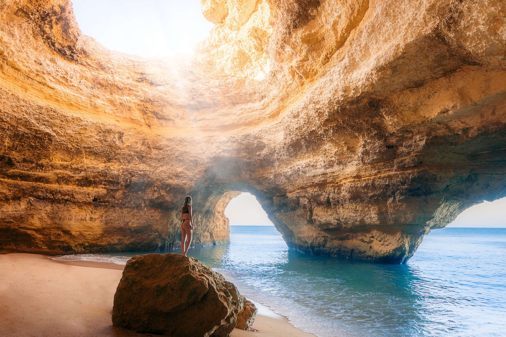 Woman standing on a rock inside Benagil Cave in Algarve, Portugal, with soft sunlight filtering through the cave’s ceiling and ocean waves entering the sea cave