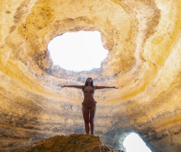 Person standing with arms outstretched inside the Benagil Cave in Algarve, Portugal, beneath the famous natural skylight in the cave ceiling